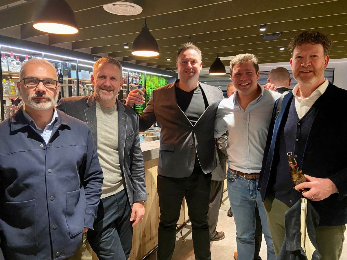 A group of five men stands together in a stylish bar. They are smiling and dressed in smart casual attire. The background features shelves filled with various drinks, and the ambiance is warm with pendant lighting.